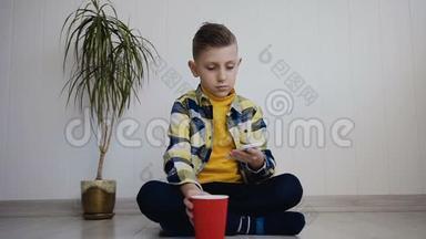 一个英俊的小男孩坐在靠近白色墙壁的地板上，用手机和红色的饮用水